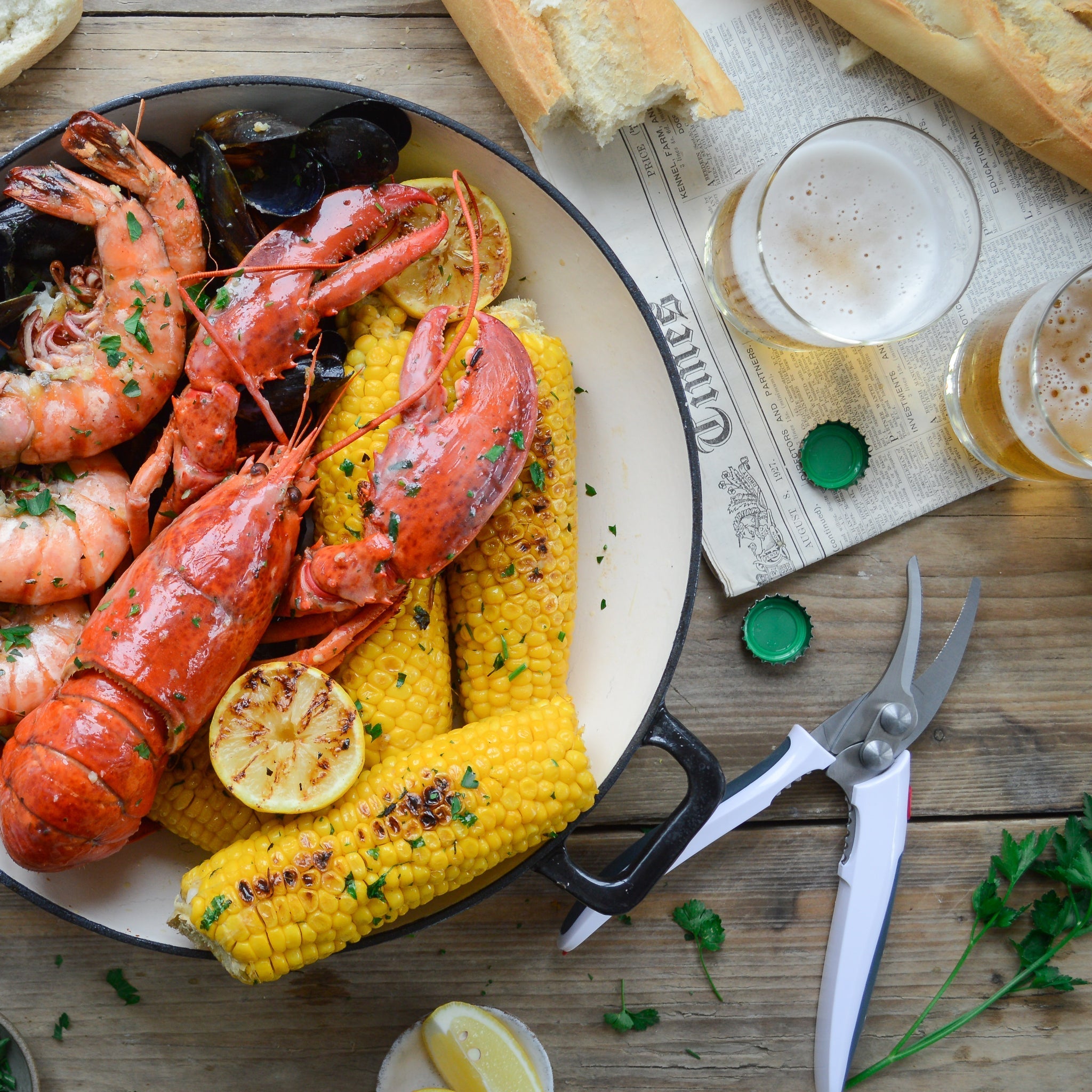 A dish with lobster, shrimp, mussels, corn on the cob, and grilled lemon sits on a wooden table with baguette, beer, herbs, and seafood shears nearby.