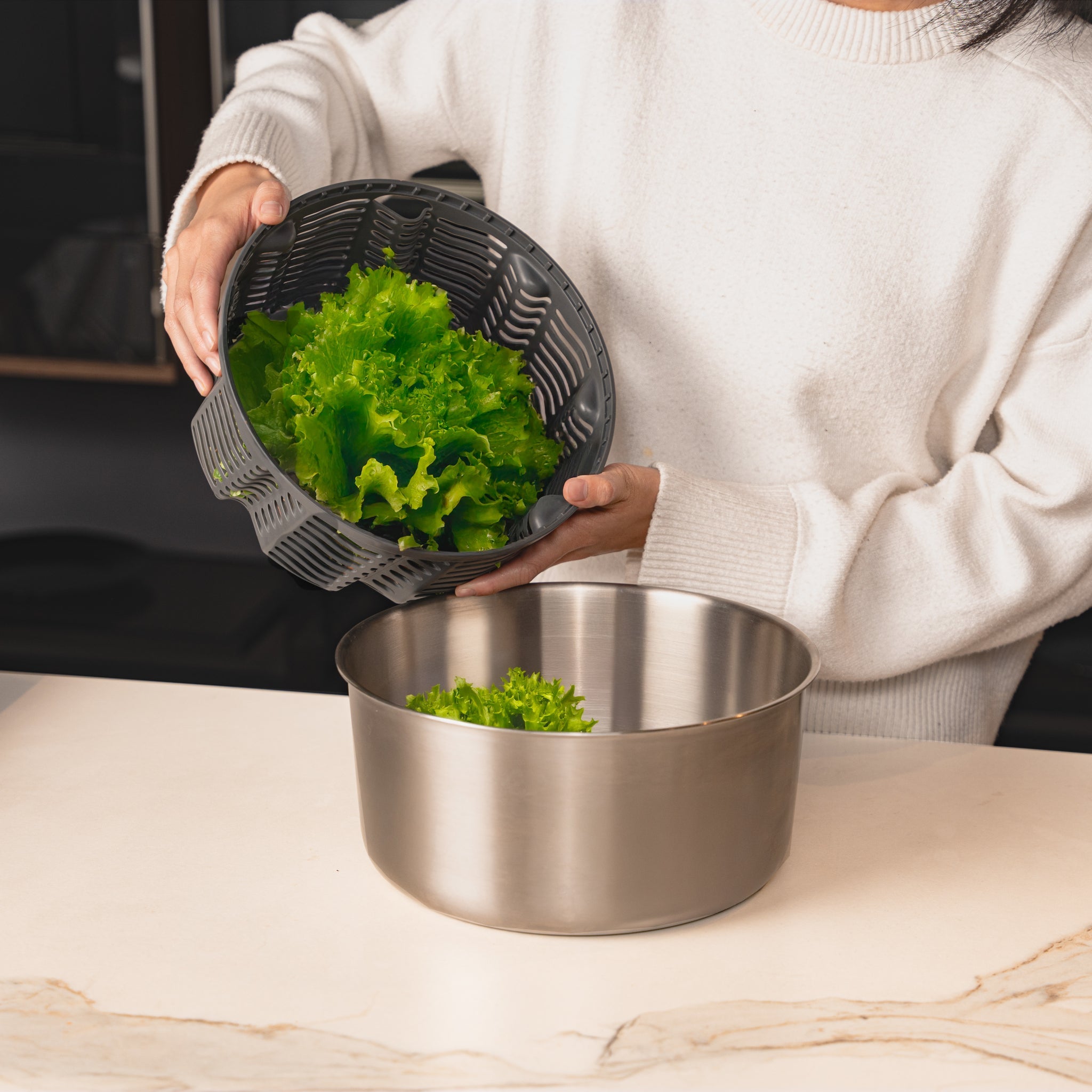 A person using a salad spinner, lifting the perforated basket filled with leafy greens over a stainless steel bowl on a kitchen counter.