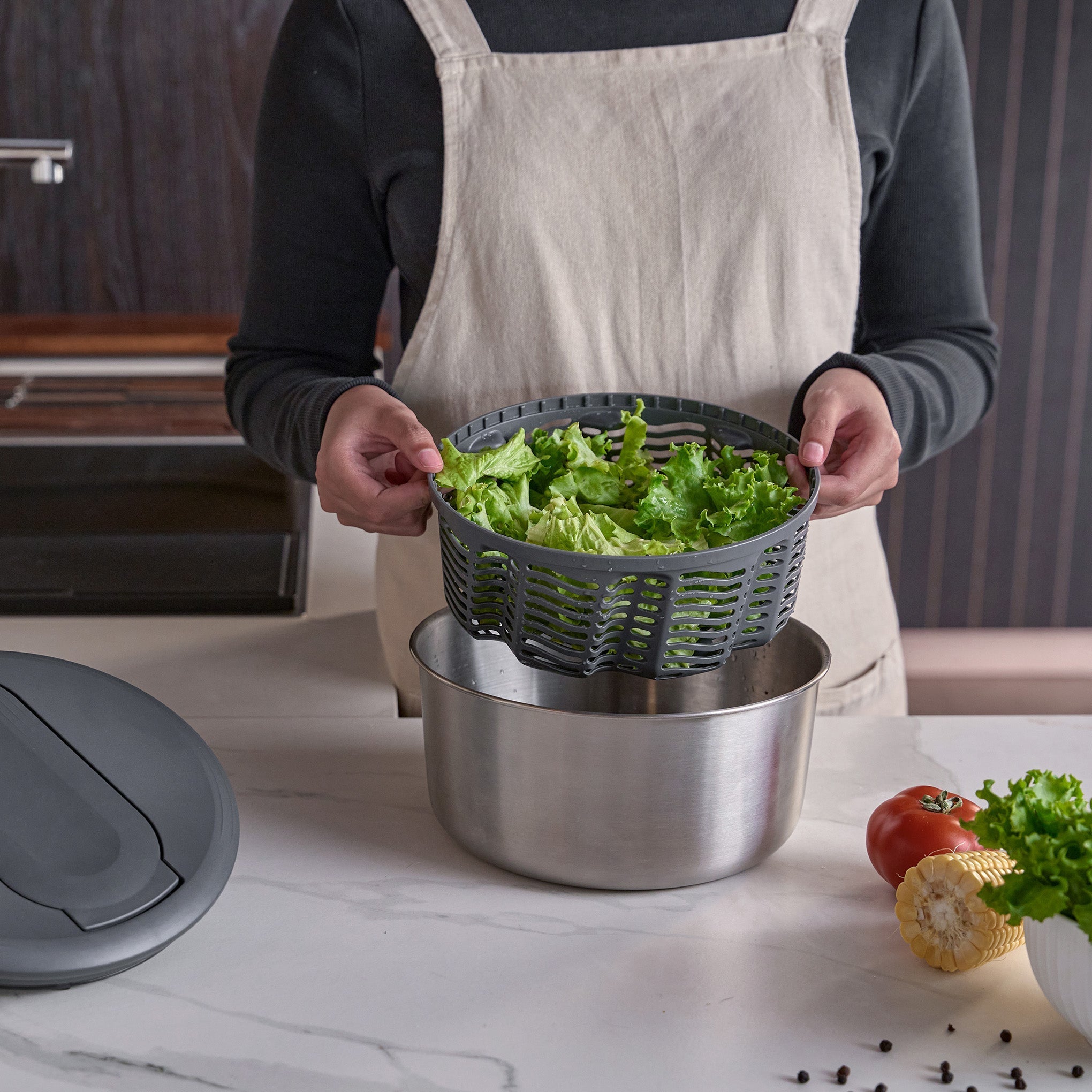 A fresh salad with greens, radishes, cucumbers, and carrots in a glass bowl, a white plate with salad, a small bowl of seeds, and a green salad spinner on a marble surface.