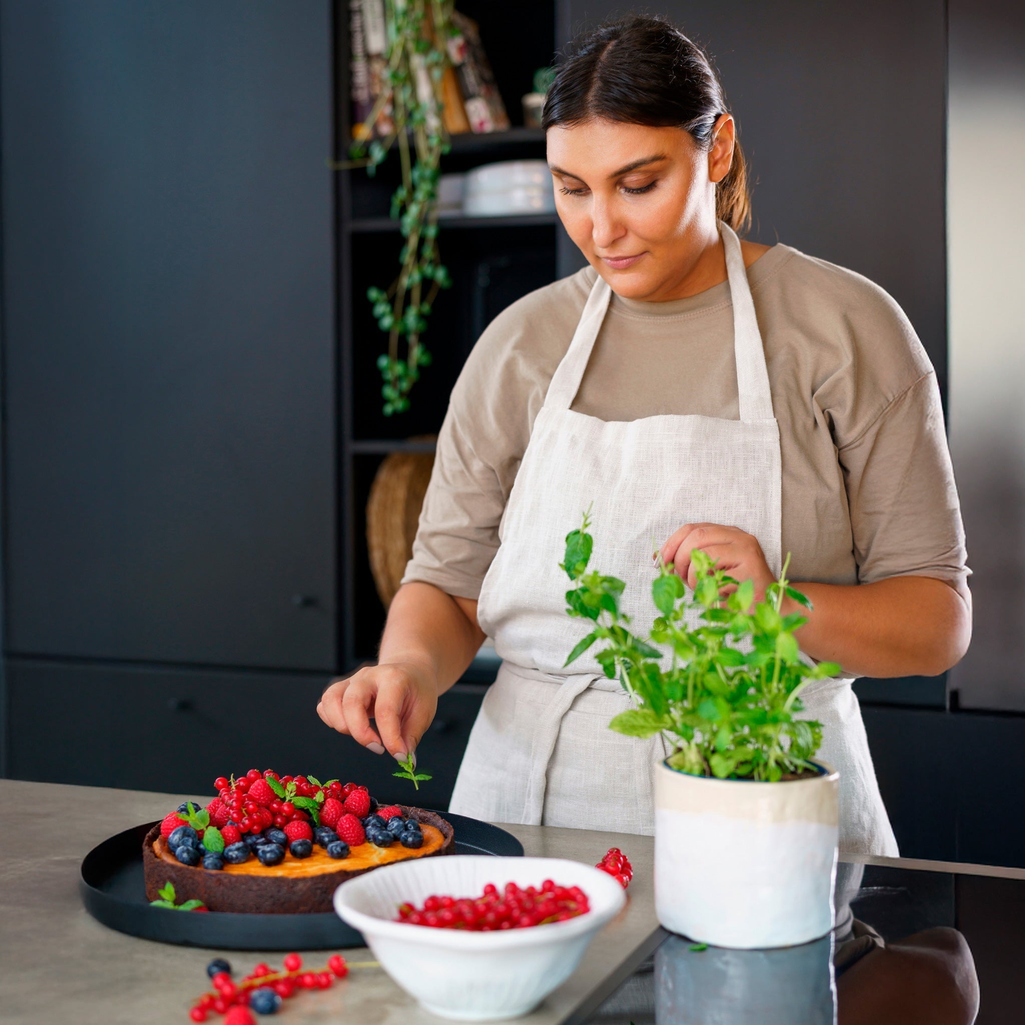 A person in a tan apron decorates a berry-topped cake with mint leaves in a kitchen. A bowl of red berries and a potted mint plant are on the counter, with dark cabinets in the background.