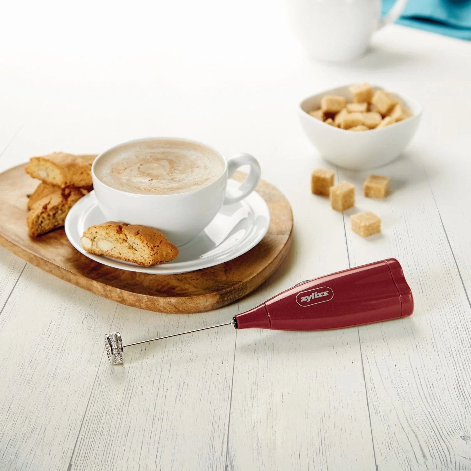 A red mug filled with coffee sits on a marble and wood table. A small red-handled spoon rests inside the mug. A plate with a fork and knife is partially visible nearby.