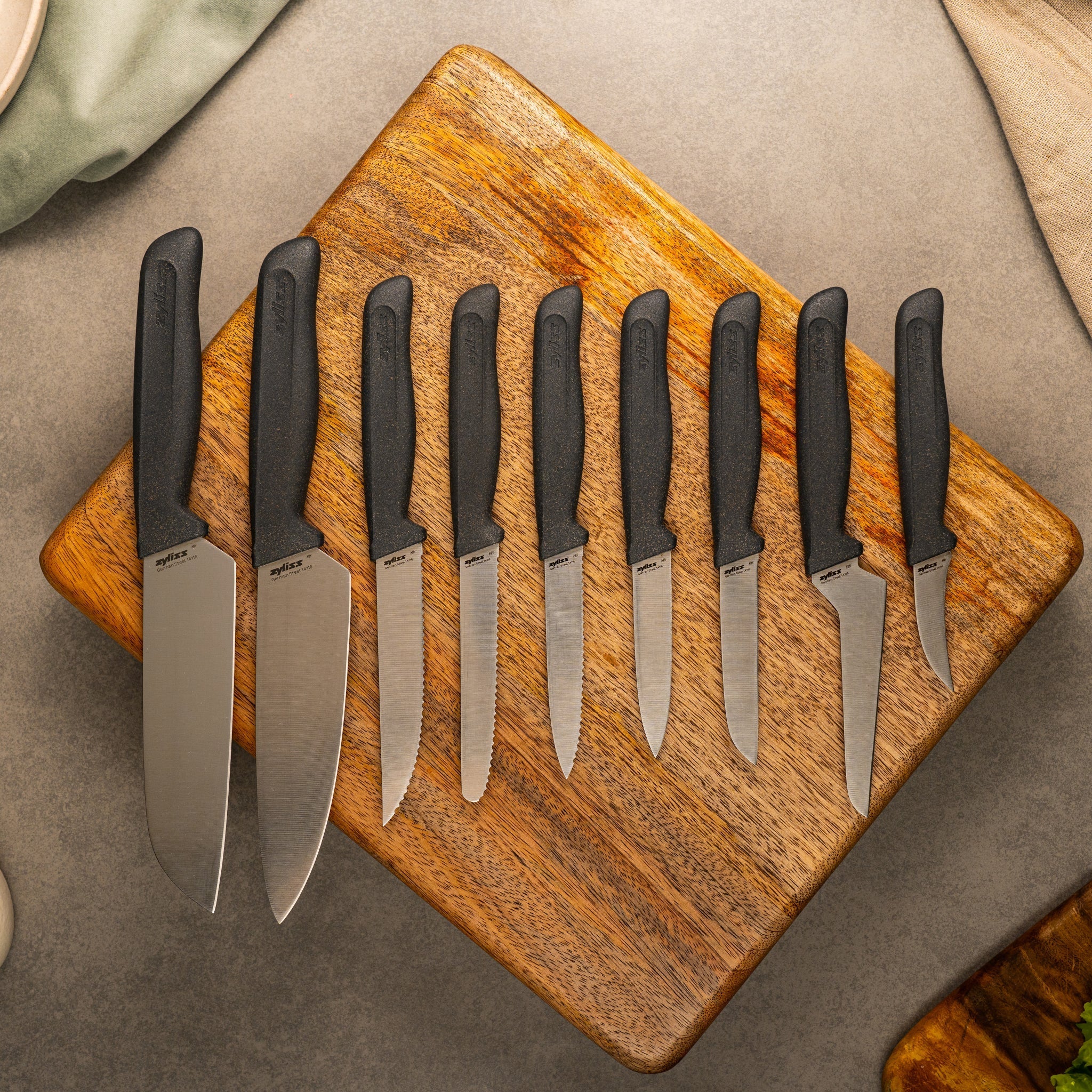 A set of seven kitchen knives with black handles displayed on a wooden cutting board. The knives vary in size and shape, suggesting different uses. The background is a neutral gray surface.