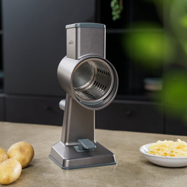 A Zyliss Gourmet Drum Grater Set with stainless steel drums stands on a kitchen counter beside whole potatoes and a plate of shredded cheese, set against a blurred dark background with hints of greenery on the side.