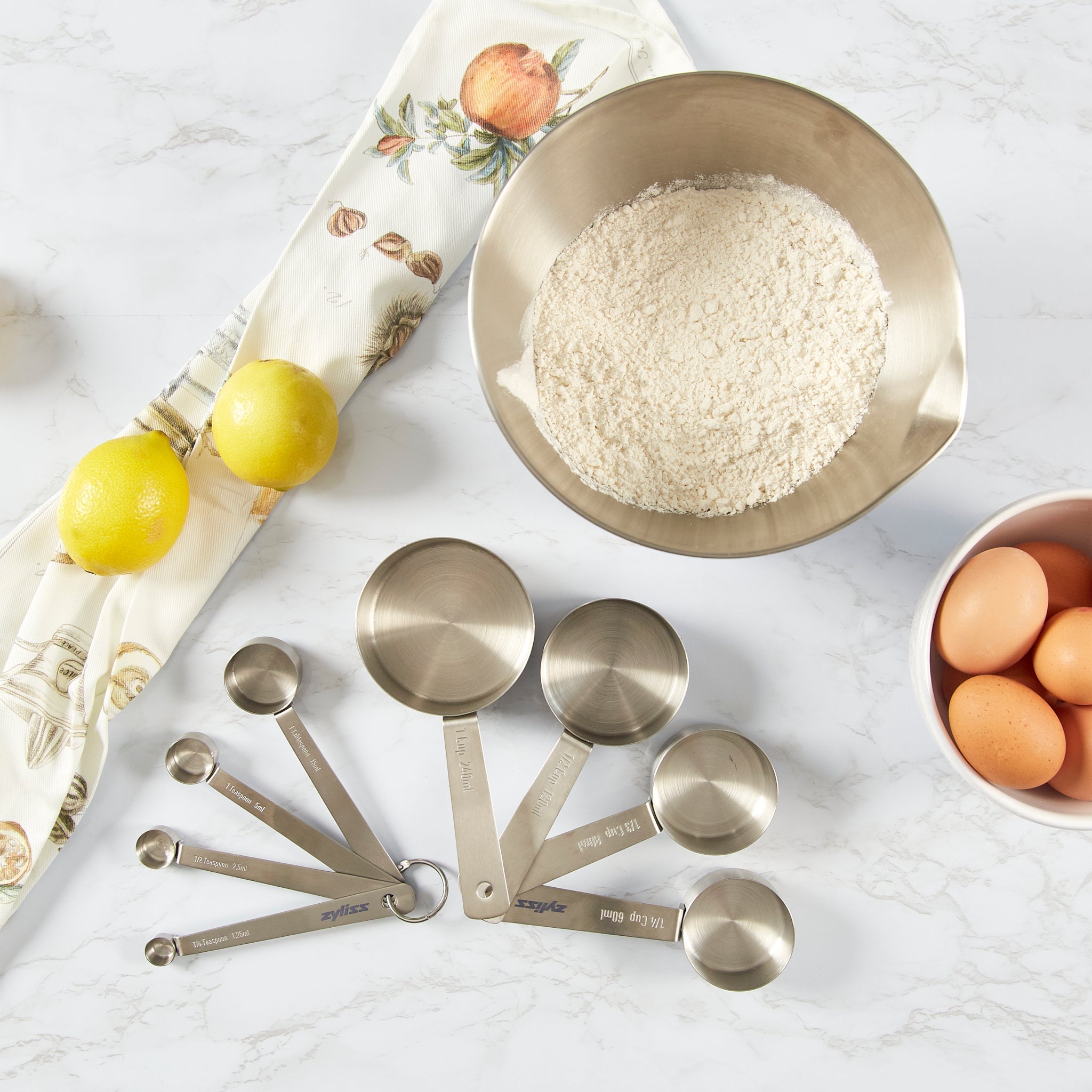 A bowl of flour, a bowl with four eggs, two lemons, Zyliss Stainless Steel Measuring Spoons, and a decorative kitchen towel are arranged on a marble countertop—perfect for any baking and cooking enthusiast.