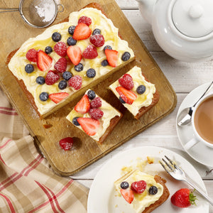 A sponge cake baked in a Zyliss Durable Non-Stick Removable Base Square Pan is topped with cream and berries, displayed on a wooden board next to a teapot, tea cup, and a plate holding a sliced piece of cake on a white table.