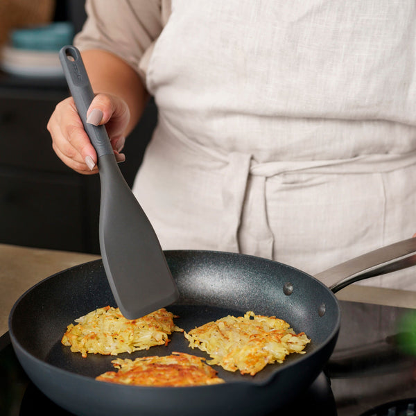 Wearing a light apron, someone uses the Zyliss Easy Lift Turner to cook three potato pancakes in a nonstick frying pan on the stove.