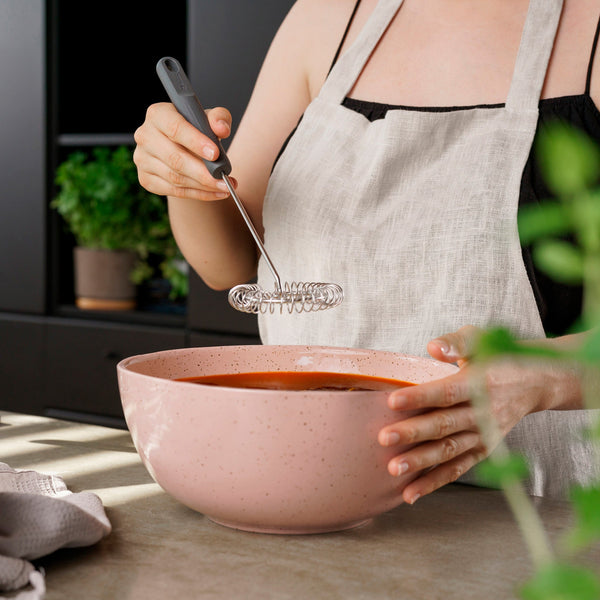 Wearing a light apron, a person uses the Zyliss Gravy Whisk to mix ingredients in a large pink bowl on the kitchen counter. Green plants and a towel appear in the background, enhancing the sauce preparation scene.