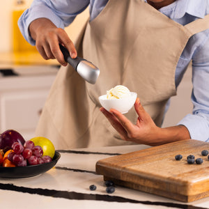 Wearing a beige apron, a person uses the Zyliss Ice Cream Scoop with an eco-friendly handle to serve vanilla ice cream into a white bowl. Nearby are assorted fruit on a plate and blueberries on a wooden cutting board.