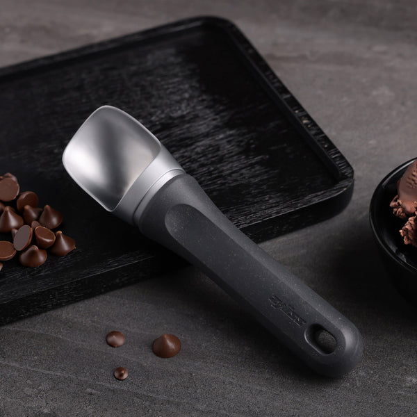 A Zyliss Ice Cream Scoop with an eco-friendly handle sits on a textured black tray beside chocolate chips and a bowl of chocolate ice cream, arranged on a gray surface.
