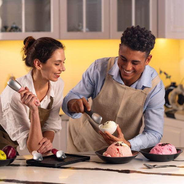 A smiling man and woman in aprons use the Zyliss Ice Cream Scoop with an eco-friendly handle to serve ice cream from bowls into a cup, enjoying dessert preparation together in the kitchen.