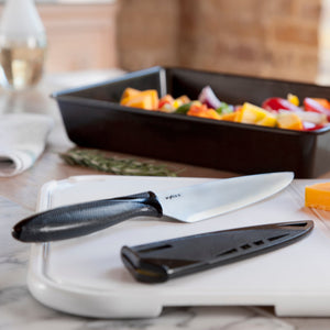 A chef’s knife from the Zyliss 6 Piece Knife Set with a black handle and sheath rests on a white cutting board beside cheese, while colorful vegetables are prepared in a baking dish in the background.