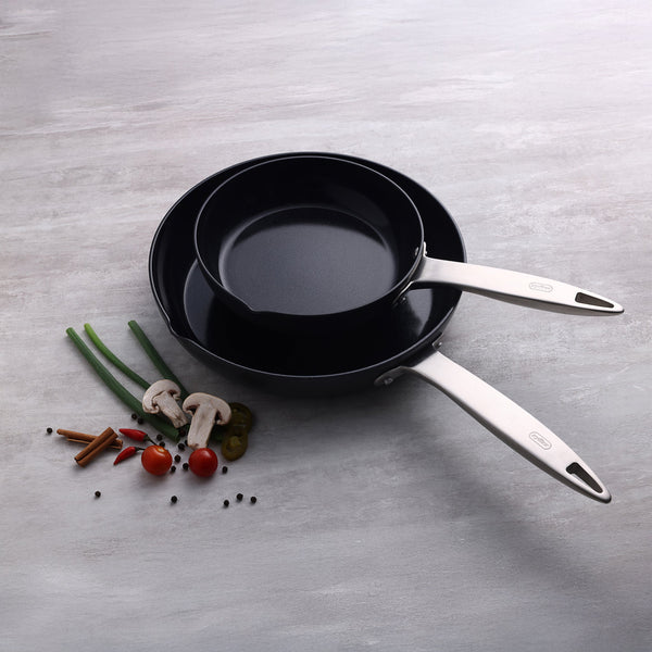 Two Zyliss Ultimate Pro Ceramic Fry Pans with silver handles are stacked on a light gray surface. Non-stick coated, they sit beside green onions, mushrooms, cherry tomatoes, green chili, cinnamon sticks, and black peppercorns. #28cm/11"