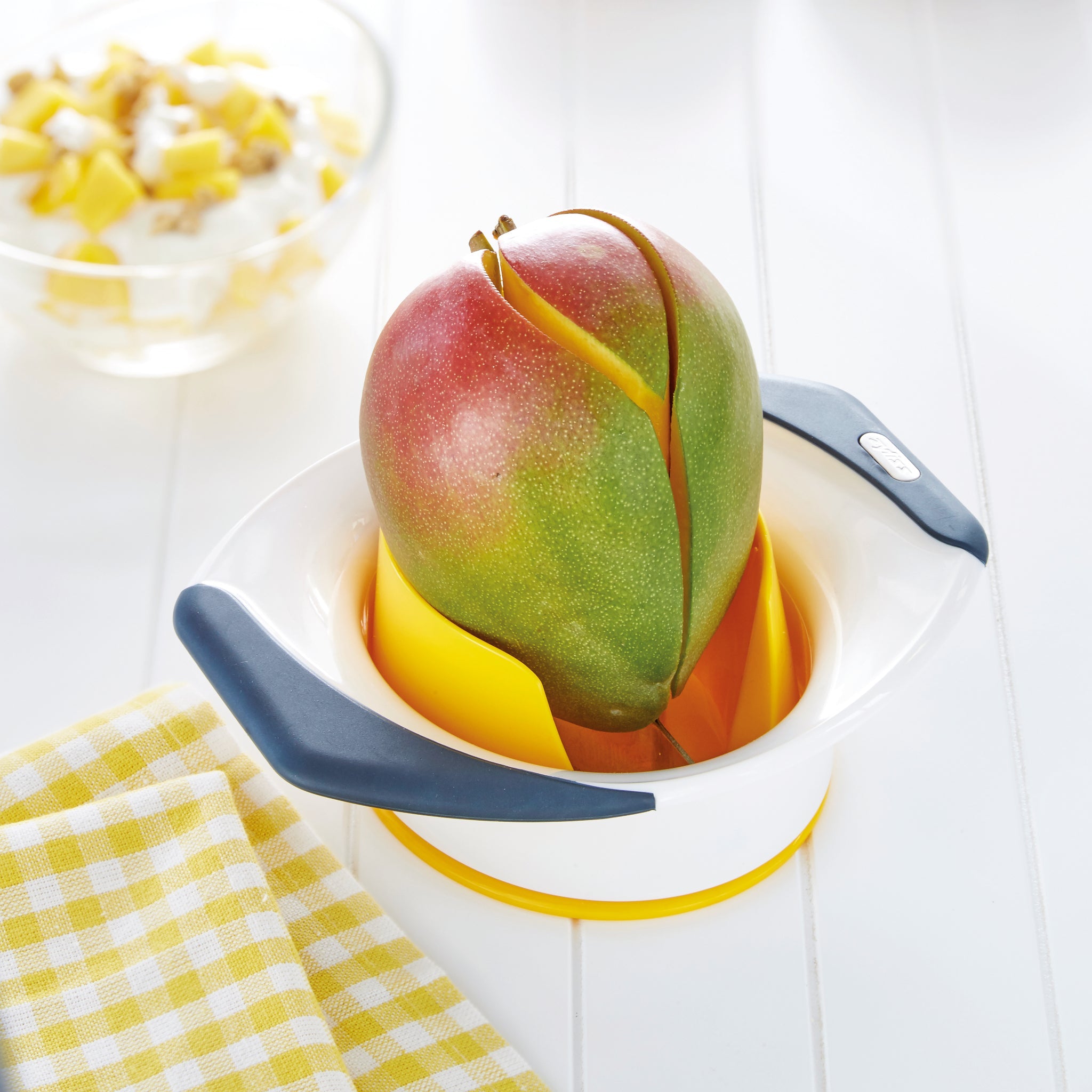 A mango is being sliced by the Zyliss Mango Splitter, featuring stainless steel blades and gray non-slip handles, on a white table next to a yellow and white checkered cloth and a bowl of diced mango pieces.