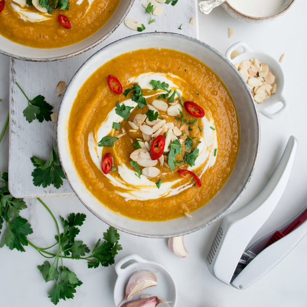 A bowl of orange soup with cream, chili, herbs, and almond flakes is surrounded by parsley, garlic, and the Zyliss Susi 3 Garlic Press featuring an ergonomic handle on a white surface.