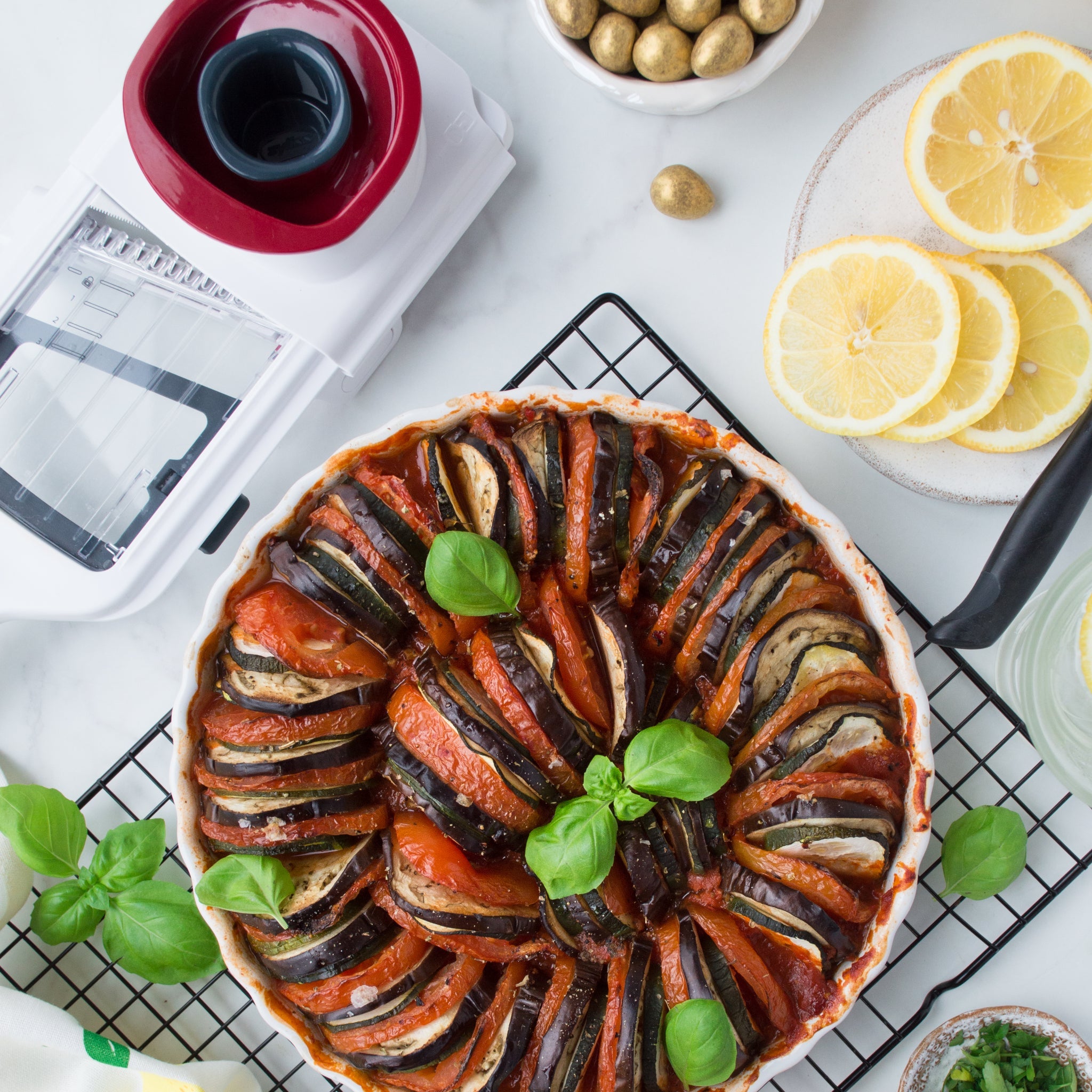 A baked ratatouille garnished with fresh basil sits on a cooling rack, surrounded by lemon slices, a bowl of olives, the Zyliss Easy Control Handheld Slicer with thickness selector, and basil leaves on a marble countertop.