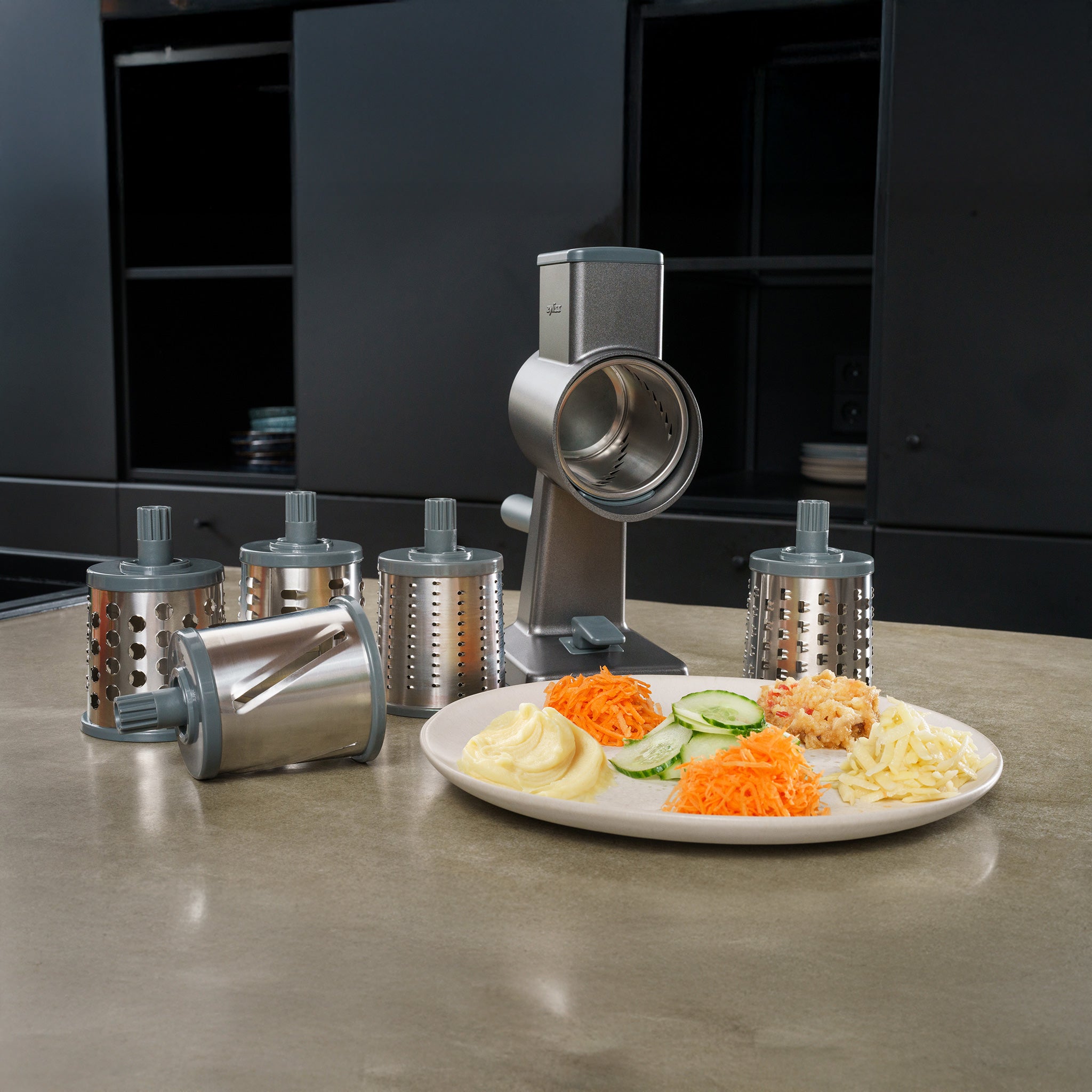 A rotary grater with five interchangeable drums is displayed on a countertop next to a plate of grated and sliced vegetables, including carrots, cucumbers, and cheese.