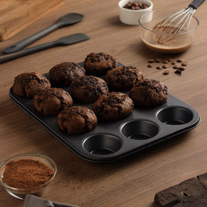 A Zyliss Non-Stick 12 Hole Muffin Pan with nine chocolate muffins rests on a wooden table, surrounded by baking tools, cocoa powder in a bowl, a whisk, chocolate chips, and a spatula.