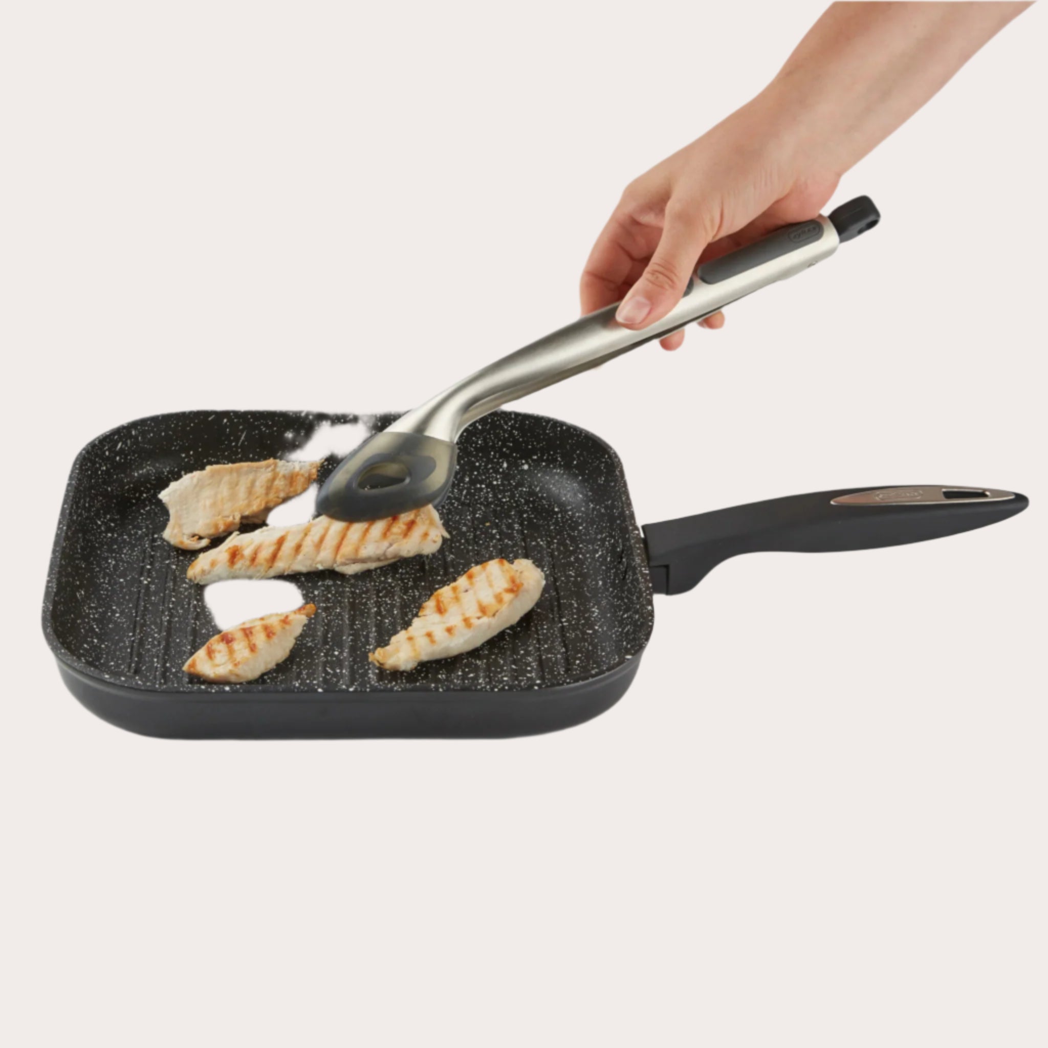 A hand uses Zyliss Silicone-Tipped Tongs to turn grilled chicken in a black, square grill pan featuring a non-stick, speckled surface.