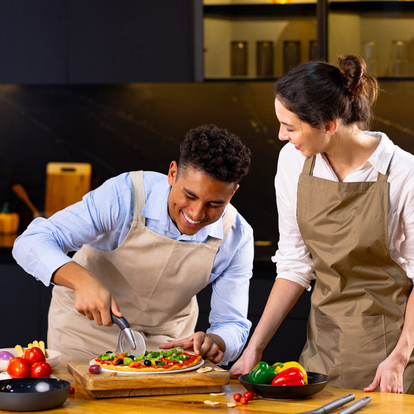 In a modern kitchen, two people in aprons smile while one slices a fresh pizza using the Zyliss Pizza & Pastry Cutter with an ergonomic handle. Colorful vegetables sit on the table, and they look like theyre enjoying cooking together. 