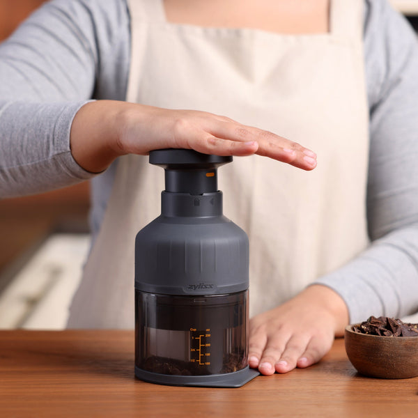 A person wearing a beige apron uses a dark gray manual food chopper on a wooden kitchen counter, with a bowl of ingredients nearby.
