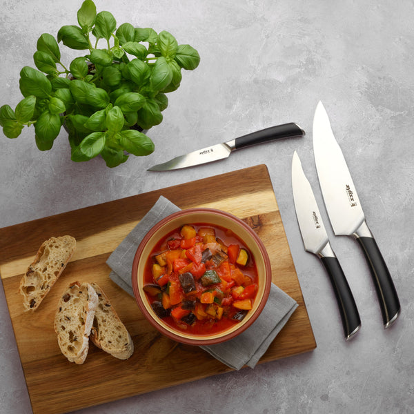 A bowl of vegetable stew on a wooden board is served with rustic bread, a napkin, and the Zyliss Comfort Pro Utility Knife 14cm / 5½, alongside a potted basil plant on a light gray surface. 