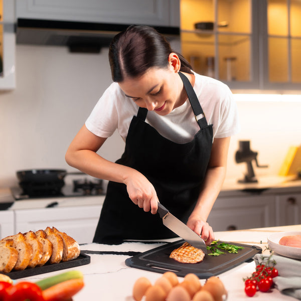 In a bright kitchen, a woman uses the Zyliss Santoku Knife 15cm / 6 with an ergonomic handle to slice cooked chicken breast on a cutting board. Sliced bread, eggs, and vegetables are seen on the nearby counter. 