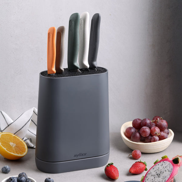 The Zyliss 6 Piece Universal Knife Block, featuring colorful, ergonomic handles, sits on a kitchen counter beside half an orange, a towel, a bowl of grapes, and sliced dragon fruit.