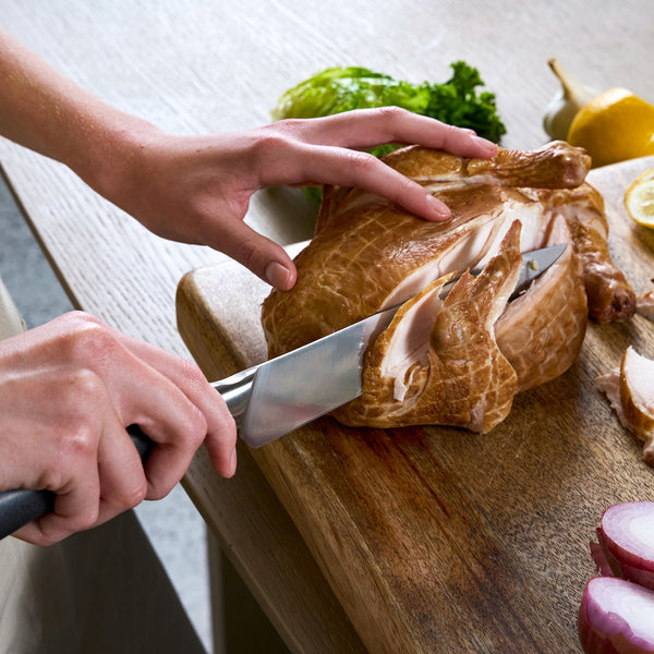 Using the Zyliss Comfort Pro Carving Knife 20cm/8", a person slices smoked chicken breast on a wooden cutting board, surrounded by fresh veggies such as lettuce, red onion, and lemon.