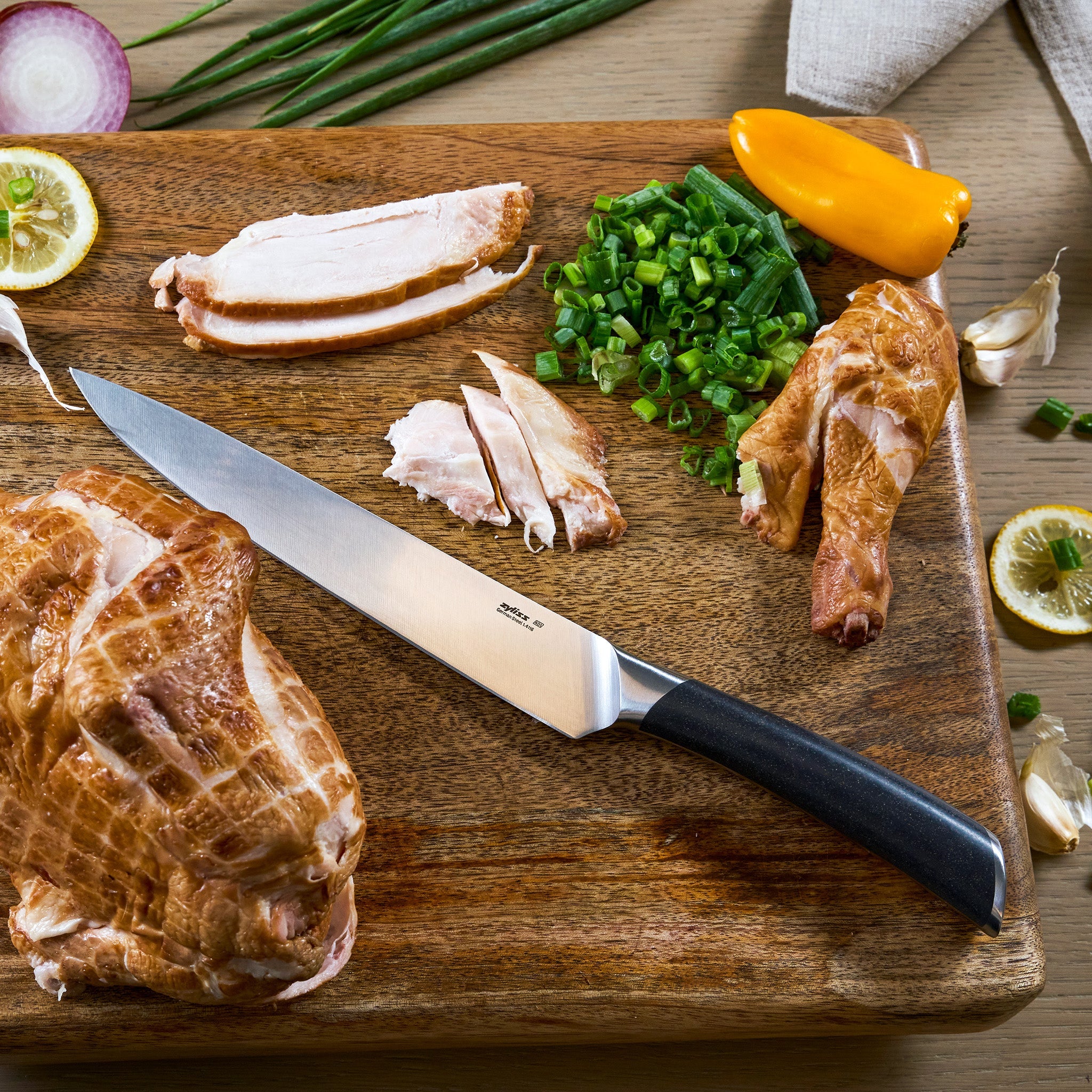 The Zyliss Comfort Pro Carving Knife 20cm rests on a wooden board with sliced smoked chicken, chopped green onions, a yellow pepper, lemon slices, garlic cloves, and green onion stalks.