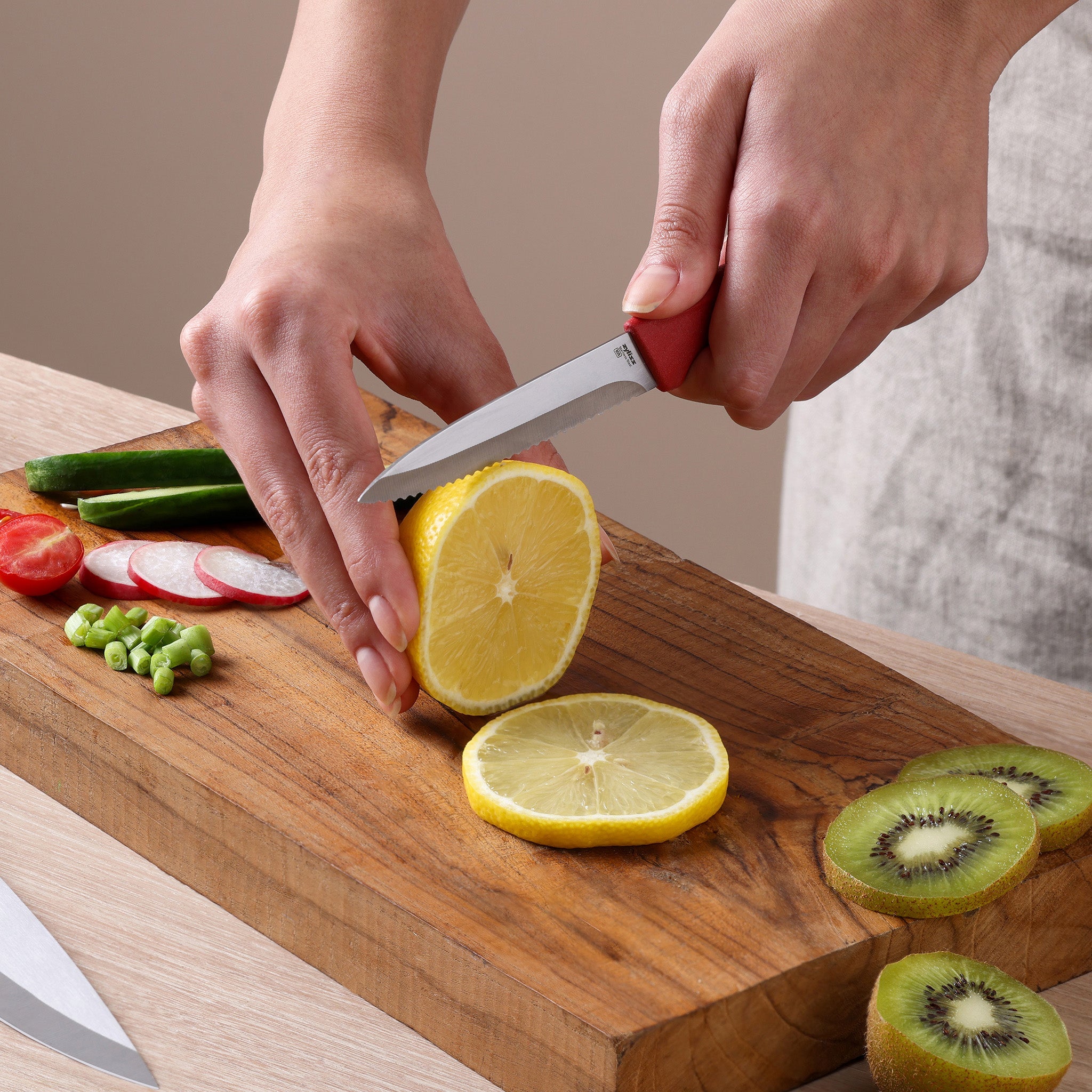 Using the Zyliss Classic 3-Piece Paring & Utility Knife Set, a person slices a lemon on a wooden cutting board surrounded by cut kiwi, cucumber, radish, tomato, and green beans.