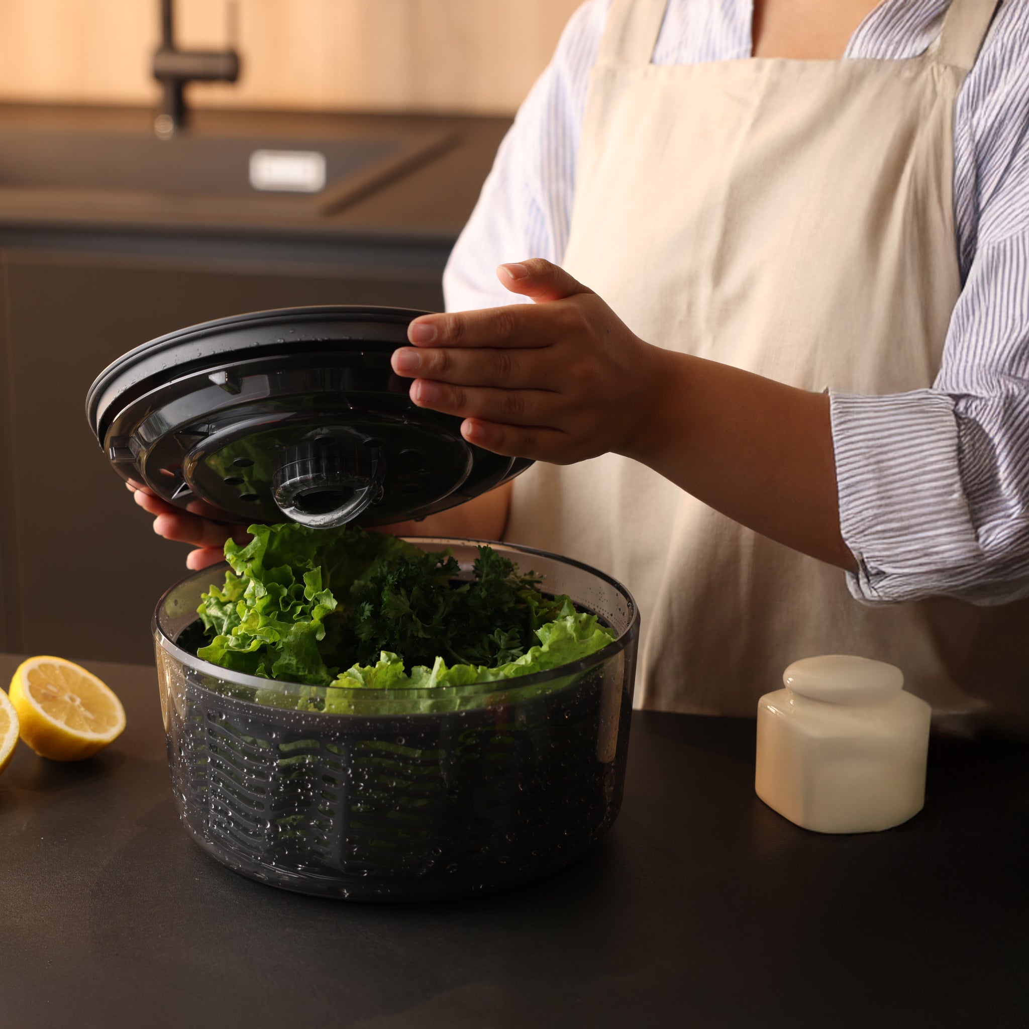 A person wearing an apron opens the lid of a salad spinner filled with fresh lettuce on a kitchen counter, with a lemon and a small container nearby.
