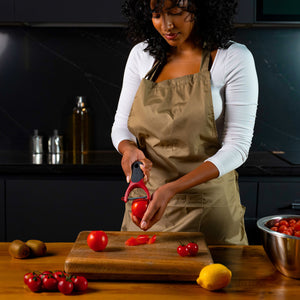 A woman in a beige apron uses the Zyliss Soft Skin Y Peeler to peel a tomato over a wooden cutting board, surrounded by more tomatoes, a lemon, kiwis, and a metal bowl of cherry tomatoes on the kitchen counter. 
