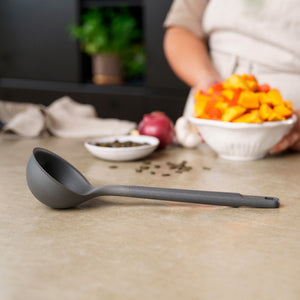 A Zyliss Ladle sits on a countertop next to a bowl of chopped orange and yellow vegetables, while a person appears blurred in the background. #Large