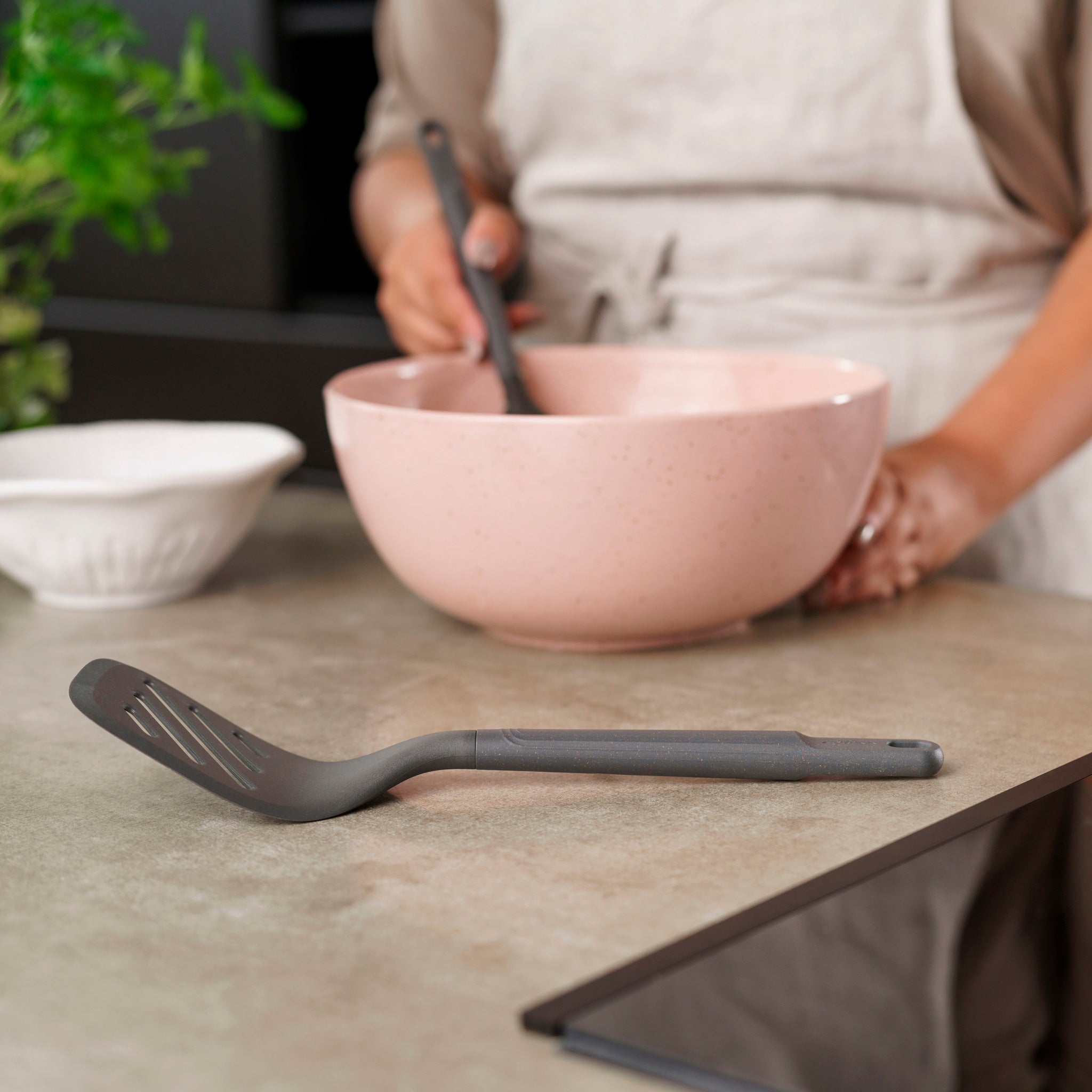 A person in an apron stirs ingredients in a pink mixing bowl, while a Zyliss Slotted Turner with an ergonomic handle rests beside a small white bowl and fresh green herbs on the kitchen counter.