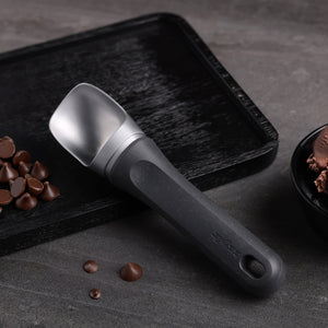 A Zyliss Ice Cream Scoop with an eco-friendly handle sits on a textured black tray beside chocolate chips and a bowl of chocolate ice cream, arranged on a gray surface. 