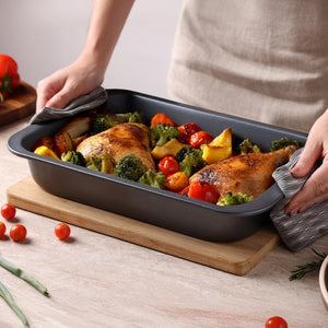 A person uses oven mitts to place a Zyliss Non-Stick Oven Tray with roasted chicken legs, broccoli, cherry tomatoes, and potatoes onto a wooden board on a light countertop.