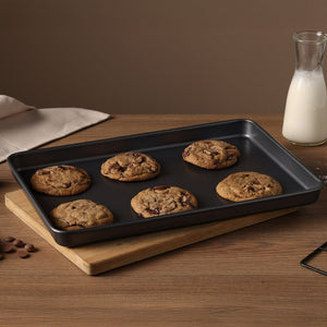 The Zyliss Non-Stick Baking Tray holds six chocolate chip cookies on a wooden board. Chocolate chips, a glass milk bottle, and a beige napkin sit nearby on the wooden table against a brown background.