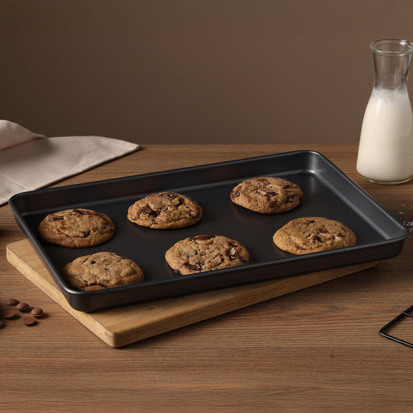 The Zyliss Non-Stick Baking Tray holds six chocolate chip cookies on a wooden board. Chocolate chips, a glass milk bottle, and a beige napkin sit nearby on the wooden table against a brown background.