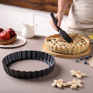 A person brushes egg wash onto a lattice-topped pie on a wooden board. Nearby is the Zyliss Non-Stick Tart Pan with removable base, flower-shaped dough, apples, blueberries, a jug, and a small bowl of egg.