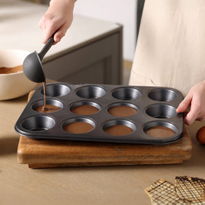 Someone pours chocolate batter into a Zyliss Non-Stick 12 Hole Muffin Pan using a ladle. The pan sits on a wooden cutting board, with waffle biscuits placed nearby on the table.